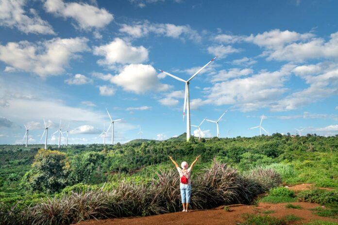 Woman Standing on a Field with Arms Raised Looking at Wind Turbines Under Blue Sky and White Clouds
